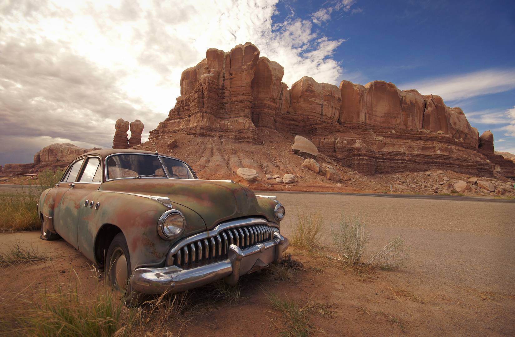 A rusted immobilized vehicle alone in the desert beside a dusty road.
